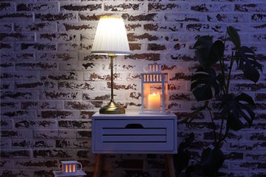 Karanlık odada tuğla duvarın yanındaki masada fenerli parlayan bir lamba.