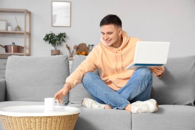 Evdeki kanepede oturan bir kupa çay ve dizüstü bilgisayarlı yakışıklı genç adam.