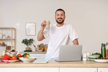 Taze sebzeli, vitaminli ve laptoplu genç adam mutfakta sağlıklı meyve suyu içiyor. Sağlıklı gıda konsepti