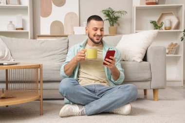 Evde cep telefonu kullanan seramik bardaklı genç adam.