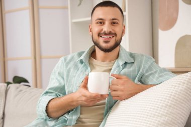 Evdeki kanepede oturan beyaz seramik bardaklı genç adam.