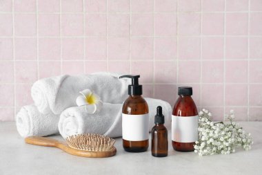 Pembe fayans duvarının yanındaki gri masada şampuan, çiçek ve havlu şişeleriyle kompozisyon