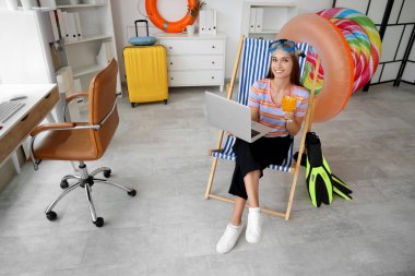 Kokteyl ve dizüstü bilgisayarlı genç iş kadını yaz tatilini ofiste bekliyor.