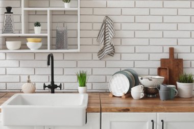 Ahşap mutfak tezgahı, lavabo, temiz tabaklar ve hafif tuğla duvarda asılı raf ünitesi.
