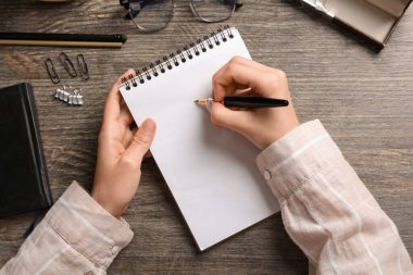 Boş defterli kadın elleri, dolma kalem, gözlük ve kırtasiye malzemeleri ahşap masada, yakın plan.