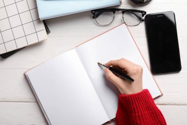 Beyaz ahşap masaya boş bir defter, gözlük ve modern cep telefonuyla bir şeyler yazan bir kadın.