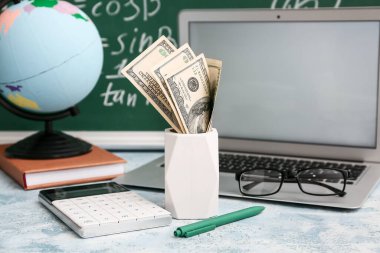 Tahtanın yanındaki masada para, hesap makinesi ve dizüstü bilgisayarlı tutucu, yakın plan. Öğrenci kredisi kavramı