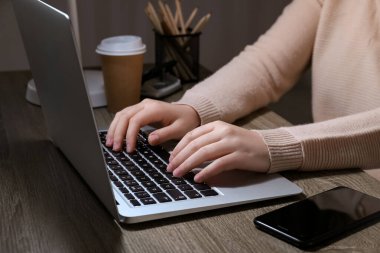 Modern dizüstü bilgisayarı, cep telefonu ve kahve bardağı olan bir kadın akşamları ahşap masada çalışıyor, yakın plan.