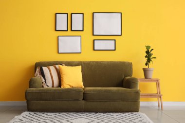 Rahat bir kanepe, ev bitkisi ve odanın sarı duvarında boş fotoğraf çerçeveleri.