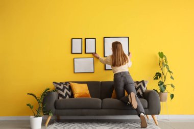 Evdeki sarı duvara boş fotoğraf çerçevesi asan bir kadın. Arka plan