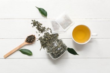 Beyaz ahşap arka planda kurumuş yapraklı bir fincan sıcak yeşil çay ve kavanoz.