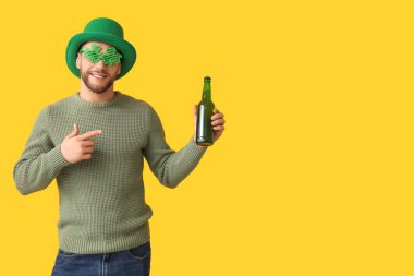 Leprikon şapkalı ve yonca şeklinde dekoratif gözlüklü genç adam sarı arka planda bira şişesini işaret ediyor. Aziz Patrick Günü kutlaması