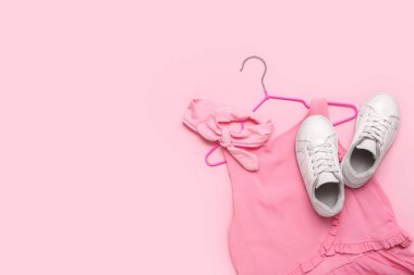 Pembe arka planda şık çocuk kıyafetleri, saç bandı ve ayakkabıları olan bir askı.