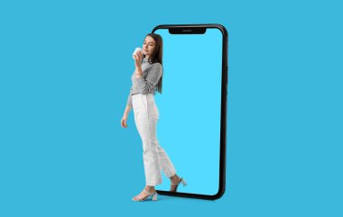 Young woman with coffee getting out of big mobile phone on blue background. Concept of digital detox clipart