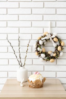Vazoda söğüt dalları, beyaz tuğla duvarda Paskalya pastası ve çelenk olan bir masa.