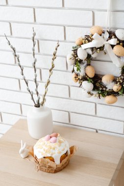 Vazoda söğüt dalları, beyaz tuğla duvarda Paskalya pastası ve çelenk olan bir masa.
