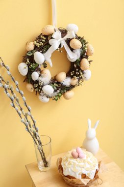 Vazoda söğüt dalları, Paskalya pastası, sarı duvarda tavşan ve çelenk olan masa.
