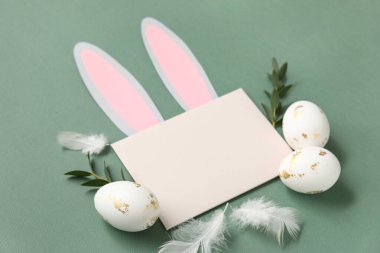 Kağıttan yapılmış tavşan kulaklı boş kart, Paskalya yumurtaları, tüyler ve yeşil arka planda yapraklar
