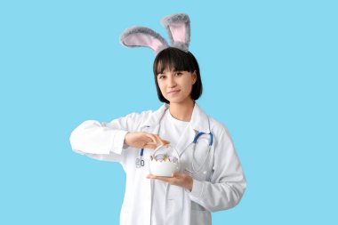 Mavi arka planda Paskalya yumurtaları olan tavşan kulaklı kadın doktor.