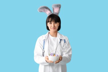 Mavi arka planda Paskalya yumurtaları olan tavşan kulaklı kadın doktor.