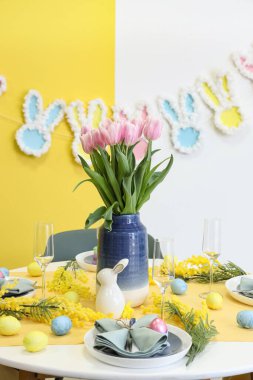 Mimoza çiçekleri, vazo lale, Paskalya tavşanı ve boyalı yumurtalarla süslenmiş şenlik masası.