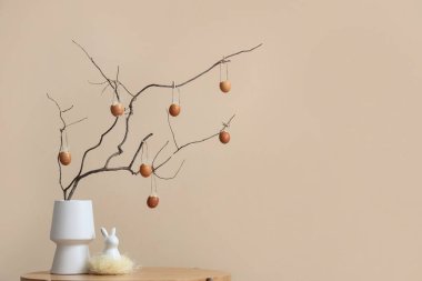 Ağaç dallı vazo, Paskalya yumurtası ve bej duvarın yanındaki masada dekoratif tavşan.