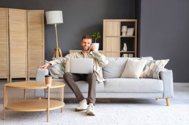 Bilgisayarlı yakışıklı adam evdeki koltukta cep telefonuyla konuşuyor.