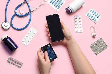 Modern cep telefonu, kredi kartı, farklı ilaçlar ve pembe arka planda steteskop kullanan kadın eller.