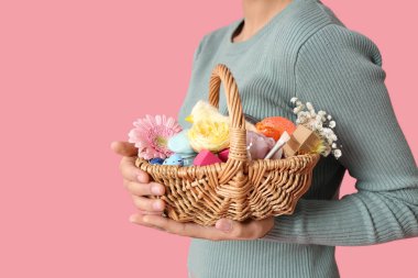 Elinde hasır Paskalya sepeti ve pembe arka planda kozmetik ürünleri tutan genç bir kadın.