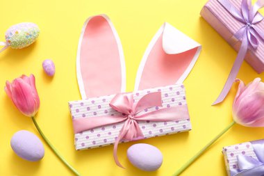 Paskalya yumurtaları, hediye kutuları ve sarı arka planda lale çiçekleri olan kağıt tavşan kulakları.