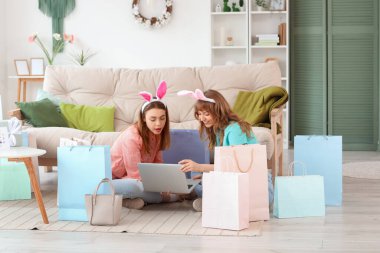 Evde dizüstü bilgisayarı ve kağıt torbaları olan tavşan kulaklı mutlu genç kadınlar. Paskalya için online alışveriş