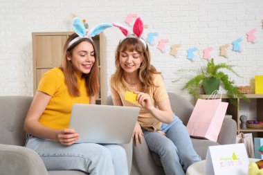 Evde dizüstü bilgisayarı ve kredi kartı olan tavşan kulaklı mutlu genç kadınlar. Paskalya konsepti için online alışveriş