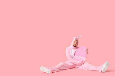 Paskalya yumurtalı tavşan kostümlü genç adam pembe arka planda oturuyor.
