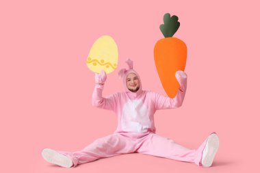 Kağıt havuç ve pembe arka planda Paskalya yumurtası olan tavşan kostümlü genç adam.
