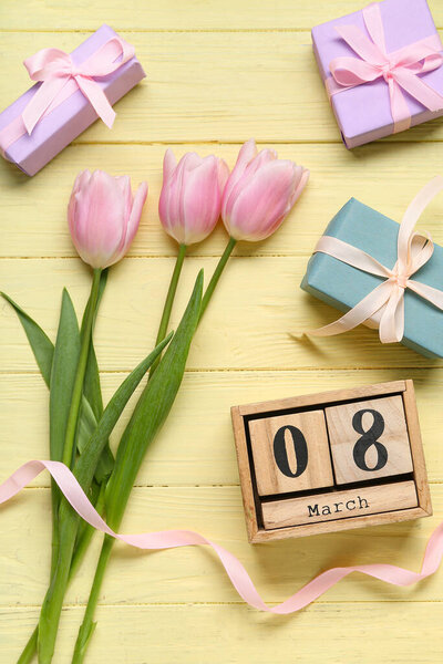 Кубический календарь с датой 8 марта, подарочные коробки и розовые тюльпаны на желтом деревянном фоне. Международный женский день. Вид сверху
