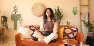 Glukofonlu genç Afro-Amerikalı kadın evdeki kanepede meditasyon yapıyor.