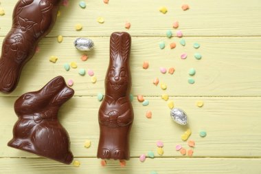 Çikolatalı tavşanlar, folyodaki Paskalya yumurtaları ve sarı ahşap arka plana serpiştirilmiş süsler.