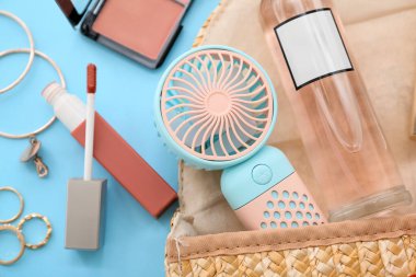 Taşınabilir elektrikli vantilatör ve mavi arka planda şık kadın aksesuarları olan kozmetik çanta.