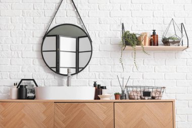 Beyaz tuğla duvarın yanında lavabo kasesi ve banyo aksesuarları olan tahta bir dolap.