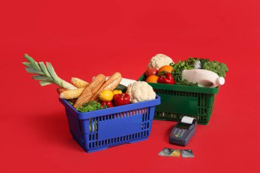 Ödeme terminali ile yiyecek dolu alışveriş sepetleri ve kırmızı arka planda kredi kartları