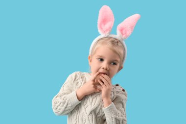 Tavşan kulaklı şirin kız mavi arka planda çikolatalı yumurta yiyor. Paskalya kutlaması