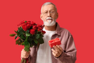 Kırmızı arka planda Sevgililer Günü için bir buket gül ve hediye taşıyan olgun bir adam.