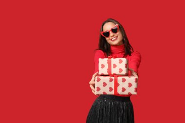 Kırmızı arka planda hediye kutuları ve güneş gözlüğü şeklinde şık bir kalbi olan güzel genç kadın. Sevgililer Günü kutlaması