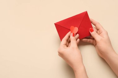 Kırmızı manikürlü kadın elleri, bej arka planda kağıt kalp ve zarf. Sevgililer Günü kutlaması