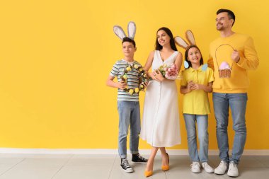 Sarı duvarın yanında Paskalya hediyeleri olan tavşan kulaklı mutlu bir aile.