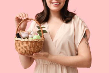 Paskalya sepetini elinde tutan pembe arka planda dekoratif kozmetik ürünleri olan genç bir kadın.