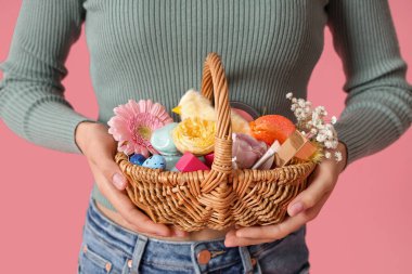 Elinde hasır Paskalya sepeti ve pembe arka planda kozmetik ürünleri tutan genç bir kadın.