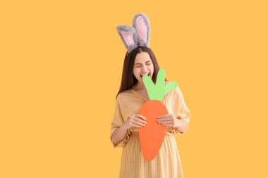 Paskalya tavşanı kulaklı, sarı arka planda kağıt havuç olan güzel, genç bir kadın.