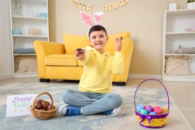 Evde paskalya için sepetli, çikolata yumurtalı ve neşeli kartpostallı tavşan kulaklı şirin bir çocuk.