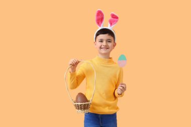 Tavşan kulaklı şirin bir çocuk. Elinde çikolata yumurtası ve turuncu arka planda Paskalya için dekorasyon sepeti var.